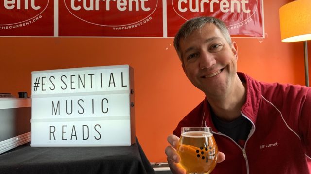 Jay holding cider in front of ESSENTIAL MUSIC READS sign
