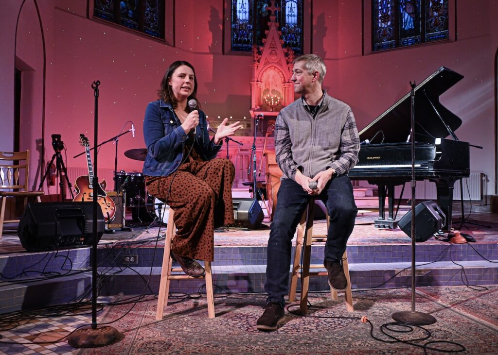 White woman in her 40s and white man of 50 sit on stools next to each other in former Catholic church. Each holds microphone; woman is speaking.