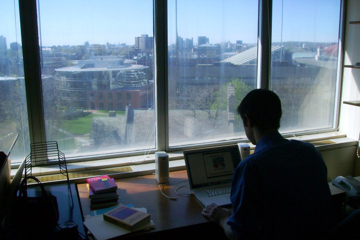 Man in silhouette types at laptop on wooden desk with books stacked atop. The Harvard University campus is visible out a window.