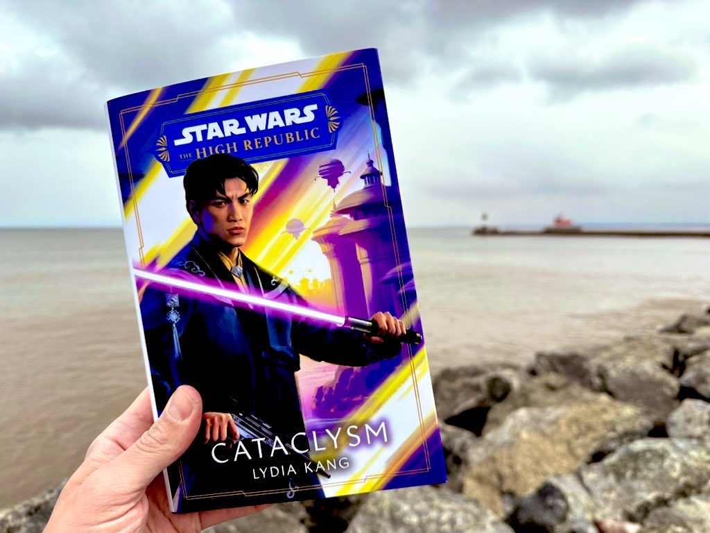 White man’s hand holding book in front of Lake Superior backdrop: “Cataclysm” by Lydia Kang with Asian man holding lightsaber on cover