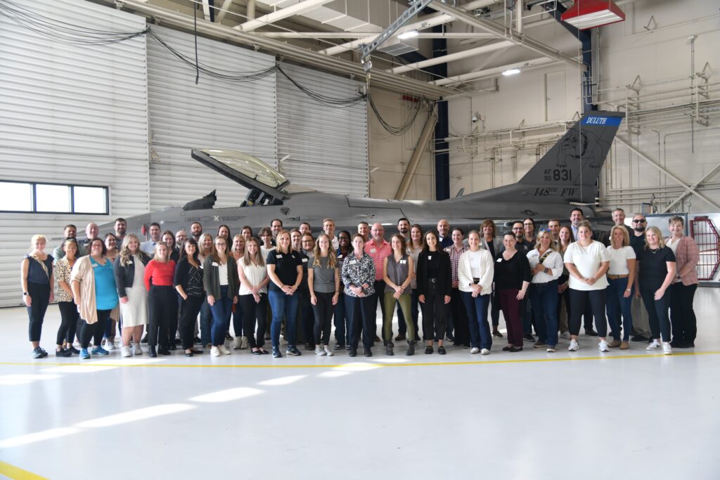 Large group of people stand in hanger in front of F-16 fighter jet