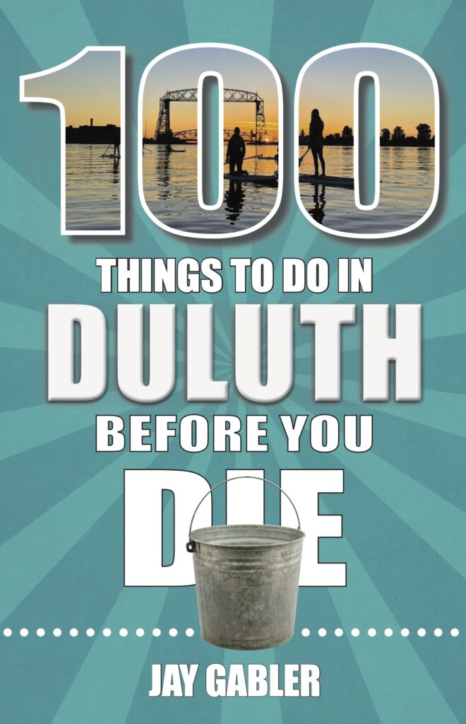 Book cover: “100 Things To Do In Duluth Before You Die” by Jay Gabler. Image of paddle-boarders near Aerial Lift Bridge is seen through “100,” and bucket is seen in front of “die.”