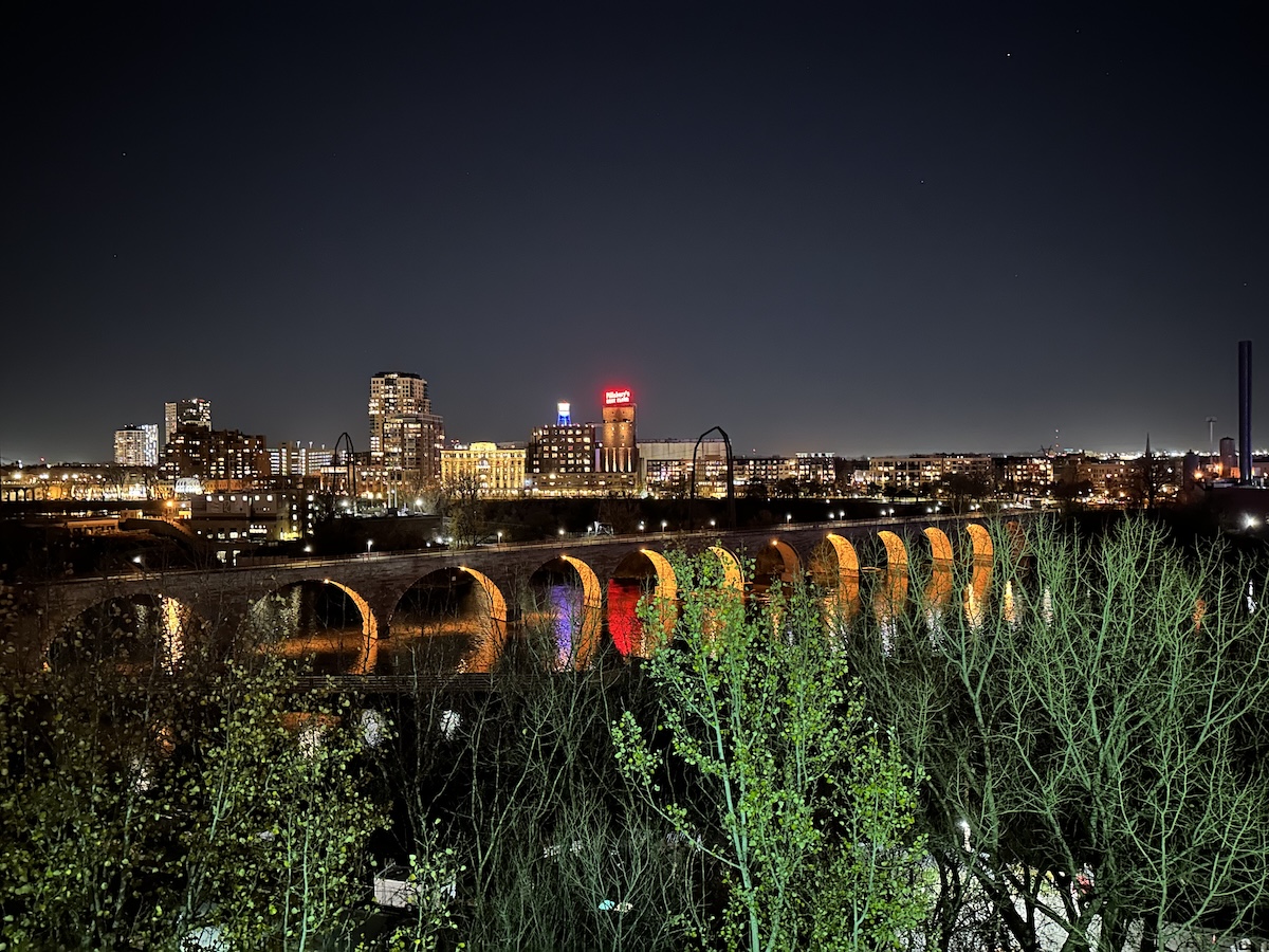 Stone bridge with arches is seen crossing a river at night in an urban setting, lit from underneath.